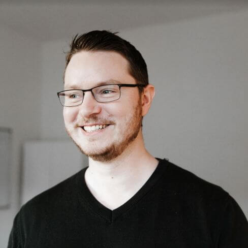Lächelnder Mann mit kurzen braunen Haaren, Brille und Bart in einem schwarzen Shirt mit V-Ausschnitt, der in einem hellen Innenbereich steht.