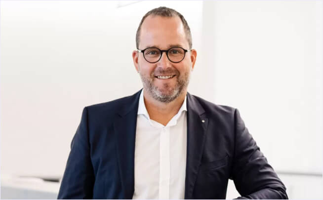Ein lächelnder, bärtiger Mann mit Brille und Anzug steht in einem hellen, modernen Büro – ideal für eine Fallstudie einer Steuerberatung.