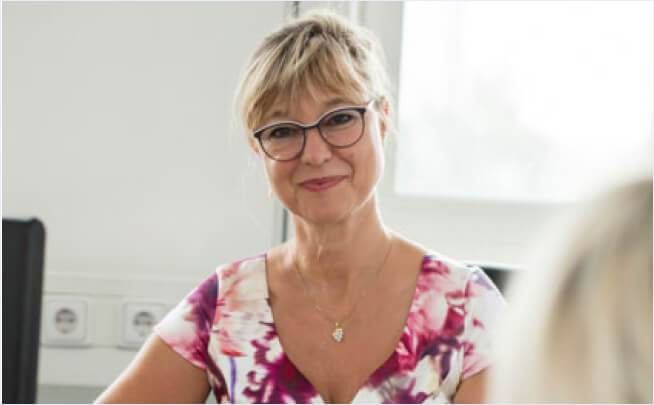 Blonde Frau mit Brille, floralem Oberteil und Herz-Kette lächelt in heller, einladender Wohnung – ideal für Immobilienanzeigen.