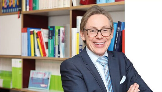Lächelnder Mann in Anzug und Brille, die Arme verschränkt, steht vor einem bunten Bücherregal und stellt einen professionellen Steuerberater dar.
