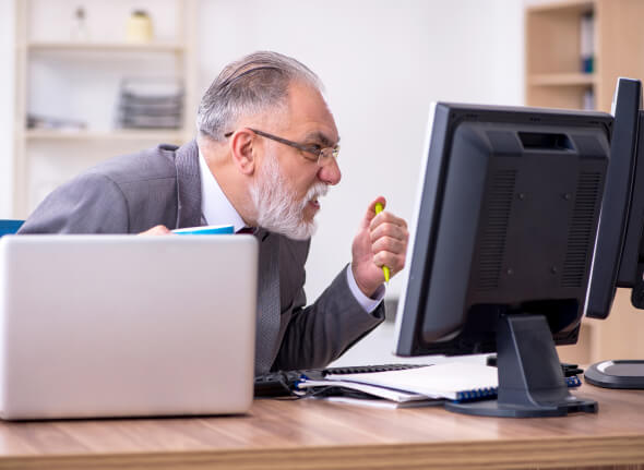Älterer Mann im Anzug ballt wütend die Faust vor dem Computerbildschirm, während er am Schreibtisch mit Laptop und Dokumenten arbeitet.