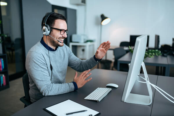 Lächelnder Mann mit Brille und Headset winkt während eines Videoanrufs an einem modernen Schreibtisch mit sichtbarem Computer, Tastatur, Maus und Zwischenablage.