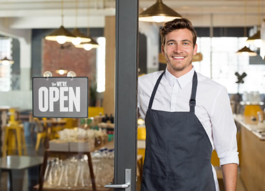 Ein lächelnder Mann in Schürze steht mit einem Schild „Ja, wir haben geöffnet“ an der Cafétür und heißt die Gäste in seinem modernen Café willkommen.
