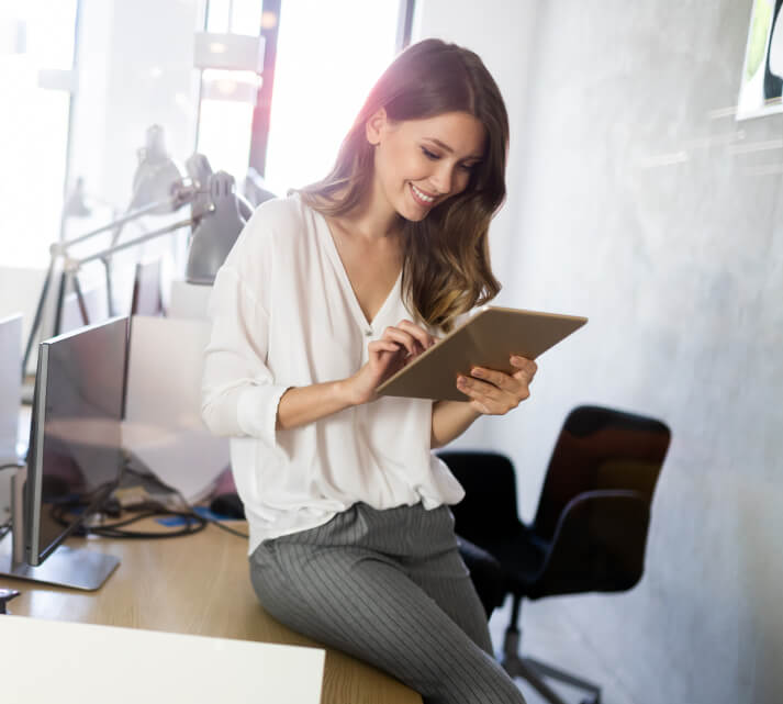 Lächelnde Frau in weißer Bluse nutzt Tablet auf dem Schreibtisch in einem modernen Local-SEO-Büro, Computer und Stühle sind sichtbar.