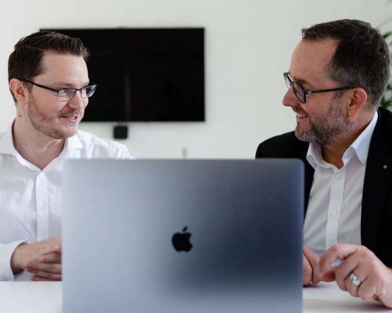 Zwei Männer mit Brillen und weißen Hemden sitzen lächelnd am Schreibtisch hinter einem offenen MacBook, TV im Hintergrund.