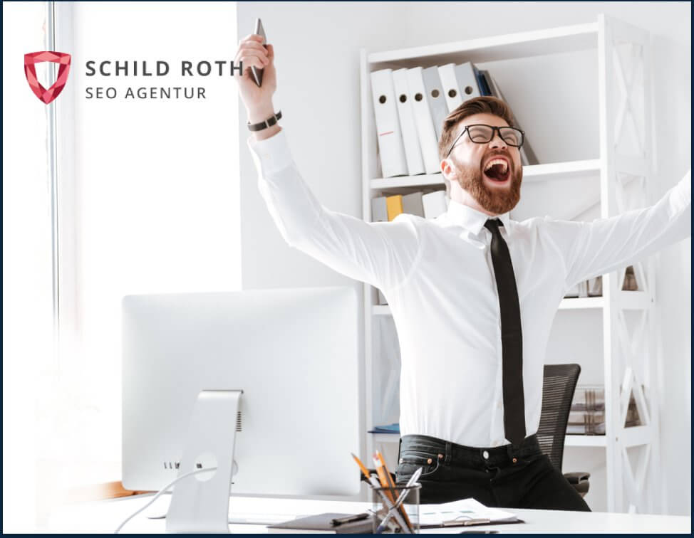 Bärtiger Mann im Business-Outfit feiert Erfolg am Schreibtisch; Logo der SCHILD ROTH SEO AGENTUR sichtbar. Helles Büroambiente.