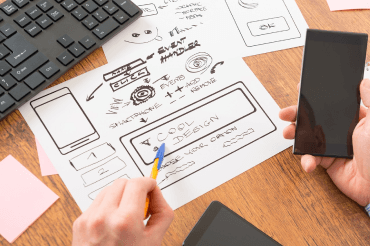 Person mit Smartphone betrachtet Website-Wireframes und SEO-Notizen am Schreibtisch, plant mobiles App- oder Website-Design.