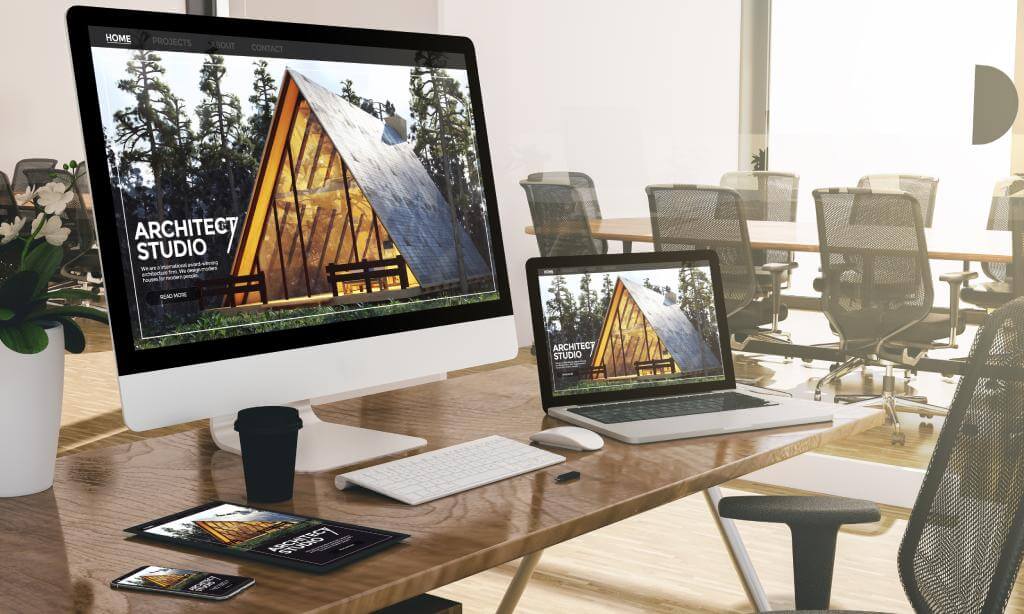 Moderner Büroschreibtisch mit mehreren Geräten, auf dem die Website eines Architekturbüros in einem hellen, offenen Arbeitsbereich mit leeren Stühlen angezeigt wird.