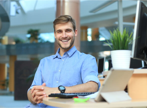 Lächelnder Mann im blauen Hemd sitzt an einem Schreibtisch mit Computer, Pflanze und Notizblock in einem modernen Büro.