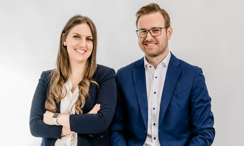 Zwei Fachkräfte der RA Büttgenbach, eine lächelnde Frau und ein lächelnder Mann in Businesskleidung, stehen selbstbewusst vor einem hellen Hintergrund.