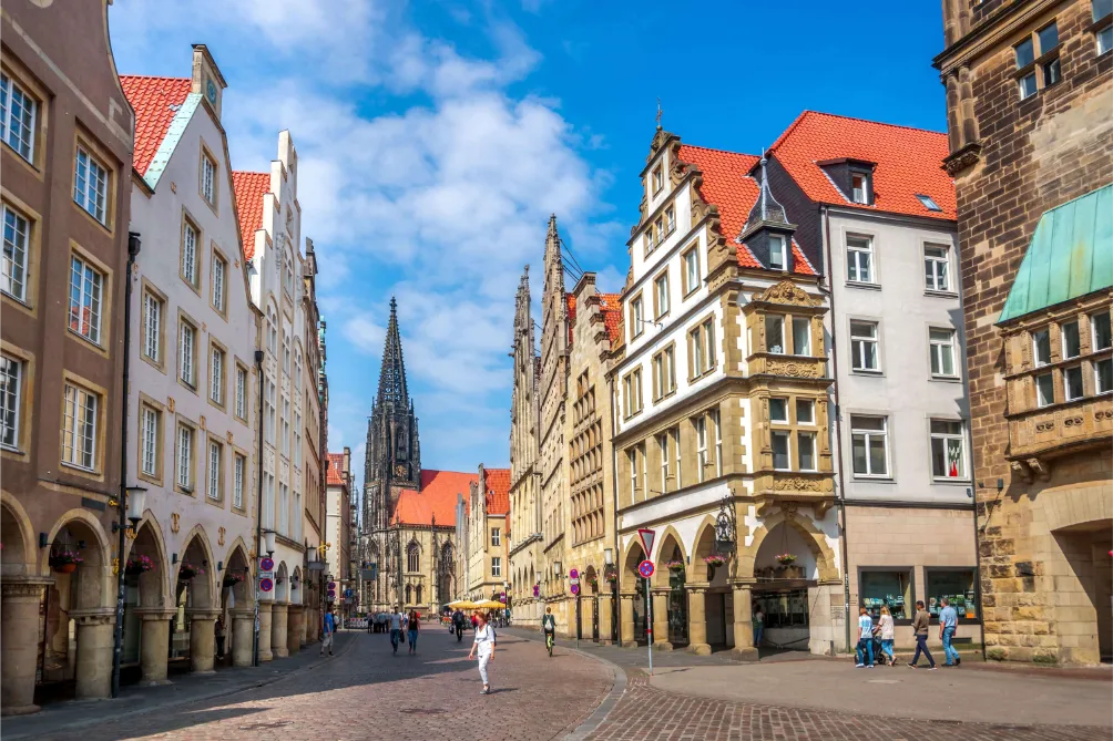 Historische Kopfsteinpflasterstraße in Münster mit Giebelhäusern, gotischer Kirche, Fußgängern und Radfahrern in der Nähe einer SEO-Agentur.