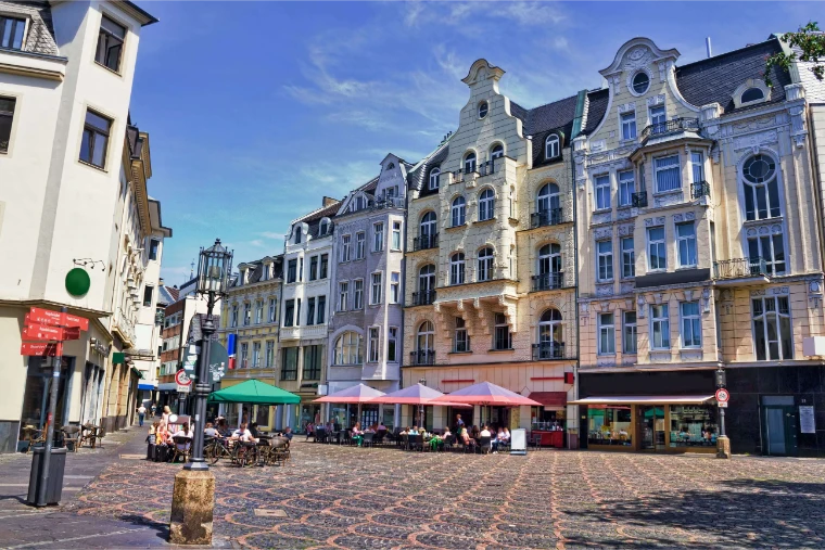 Historischer europäischer Platz mit kunstvollen Gebäuden, Straßencafés, bunten Sonnenschirmen, speisenden Menschen und gemustertem Kopfsteinpflaster unter blauem Himmel.