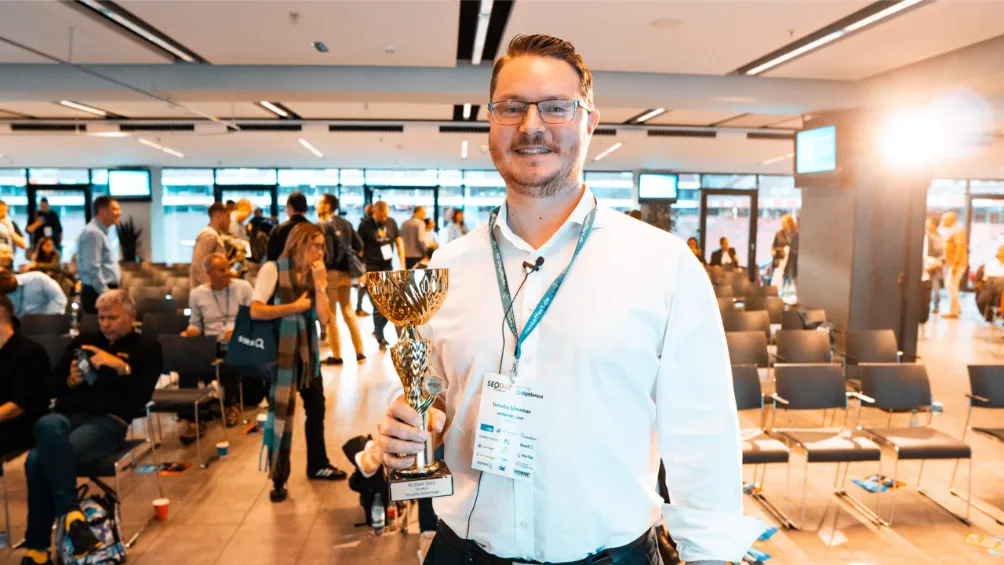 Mann im weißen Hemd mit Brille und Konferenzausweis lächelt und hält eine Trophäe; Menschen sitzen und unterhalten sich in einem modernen Veranstaltungsraum.