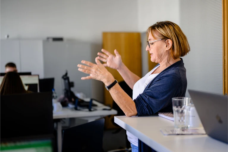 Eine Frau mit Brille spricht und gestikuliert an einem Schreibtisch mit Laptop; andere arbeiten im Bürohintergrund an Computern.