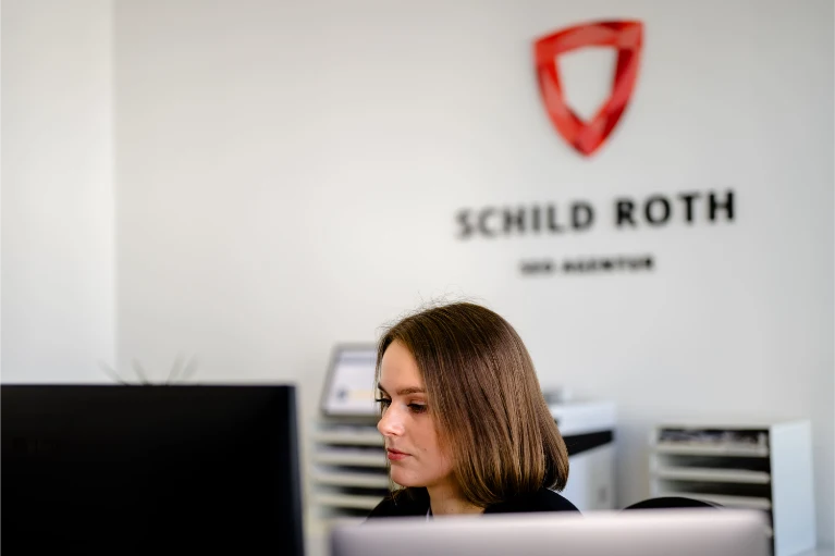 Frau mit schulterlangem braunem Haar arbeitet am Schreibtisch; an der weißen Bürowand hinter ihr sind das Schild Roth-Logo und der Agenturname zu sehen.