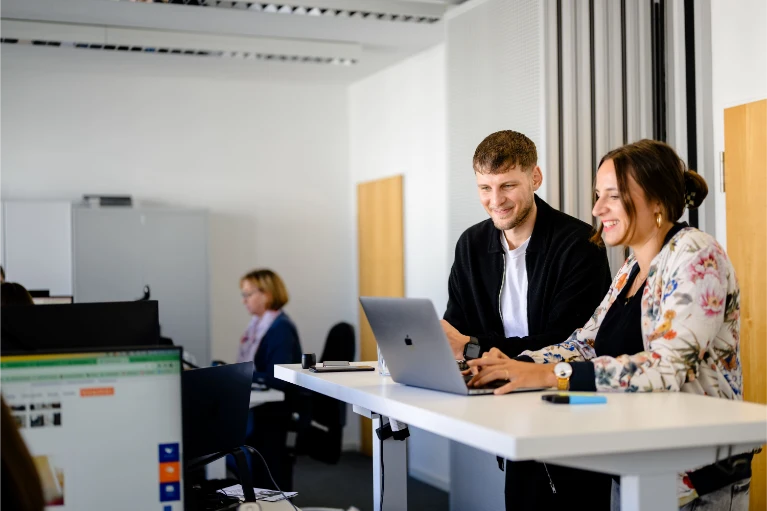 Zwei Kollegen lächeln und arbeiten gemeinsam an einem Laptop auf einem Stehpult in einem modernen Büro; eine weitere Person arbeitet hinter ihnen.