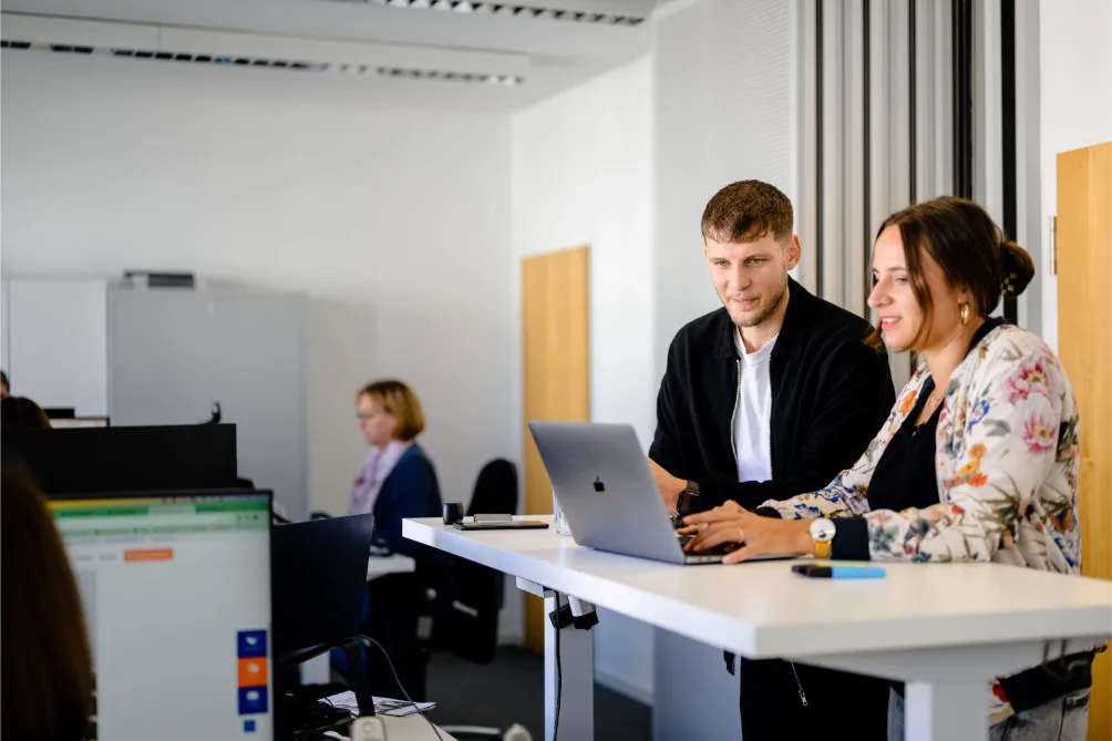 Zwei Personen lächeln, während sie an einem Laptop in einem modernen, kollaborativen Büro über SEO-Betreuung diskutieren, während Kollegen im Hintergrund arbeiten.