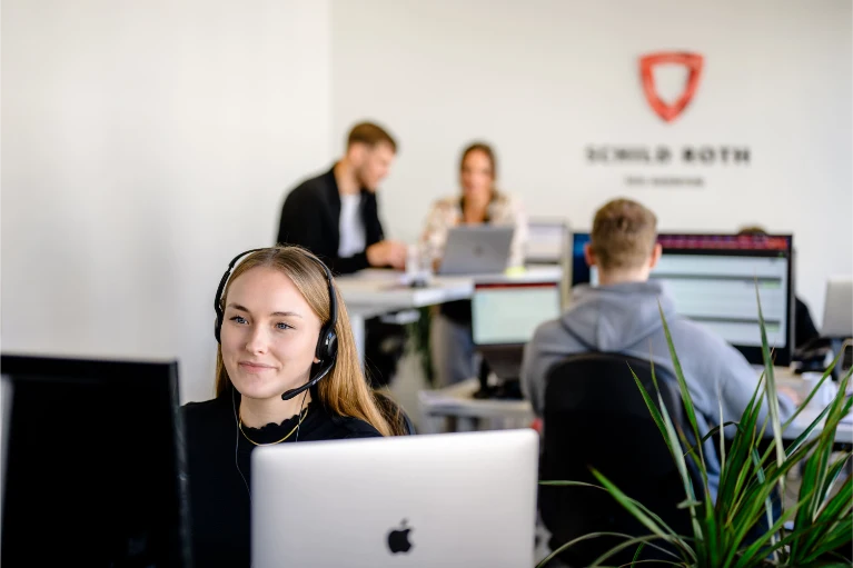 Eine lächelnde Frau mit Headset arbeitet an einem Computer; drei Kollegen arbeiten in einem hellen, modernen Bürohintergrund zusammen.