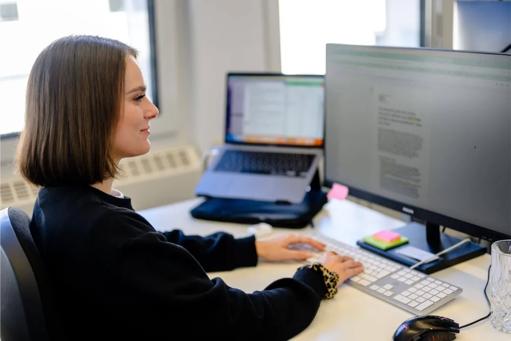 Frau mit braunen Haaren arbeitet an der SEO-Betreuung an einem Schreibtisch mit Laptop, Büromaterial und einem großen Monitor.