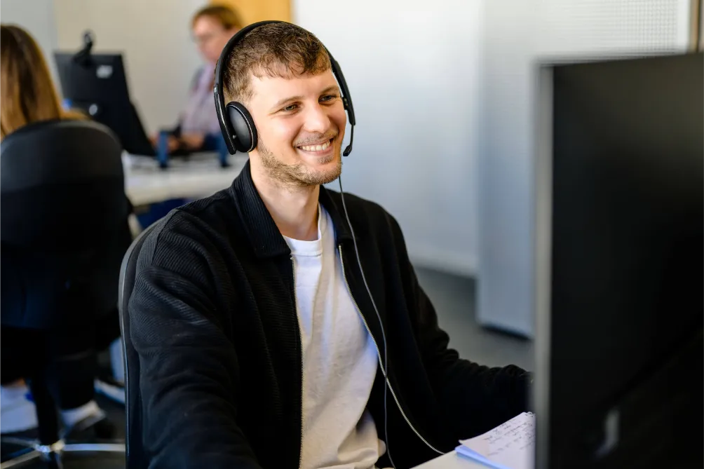 Lächelnder Mann mit Kopfhörern am Schreibtisch, schaut auf den Monitor im Büro und bespricht möglicherweise ein SEO-Konzept mit Kollegen.