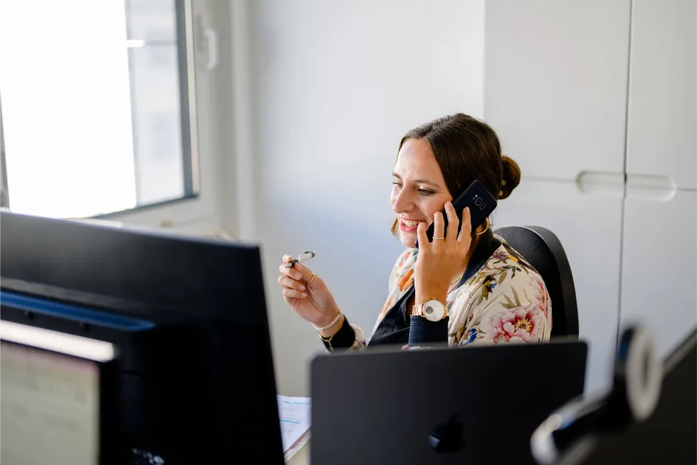 Frau am Büroschreibtisch, lächelt am Telefon, hält eine Brille; auf Computerbildschirmen wird ein SEO-Konzept angezeigt, Büromaterial liegt herum.