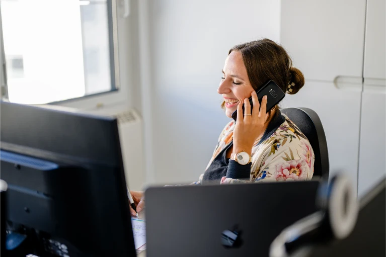 Lächelnde Frau in geblümter Jacke telefoniert an ihrem Schreibtisch vor einem Computermonitor in einem Büro.
