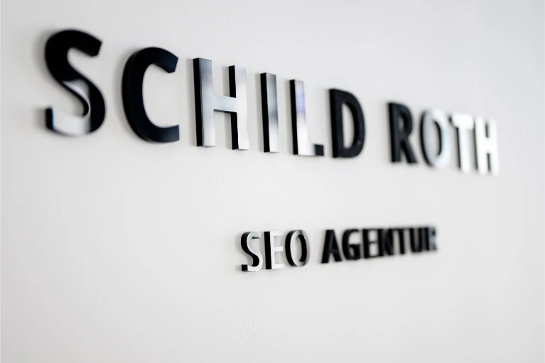 Schwarze 3D-Buchstaben „SCHILD ROTH SEO AGENTUR“ auf einer weißen Wand, rechts mit Schatten und leichter Unschärfe.