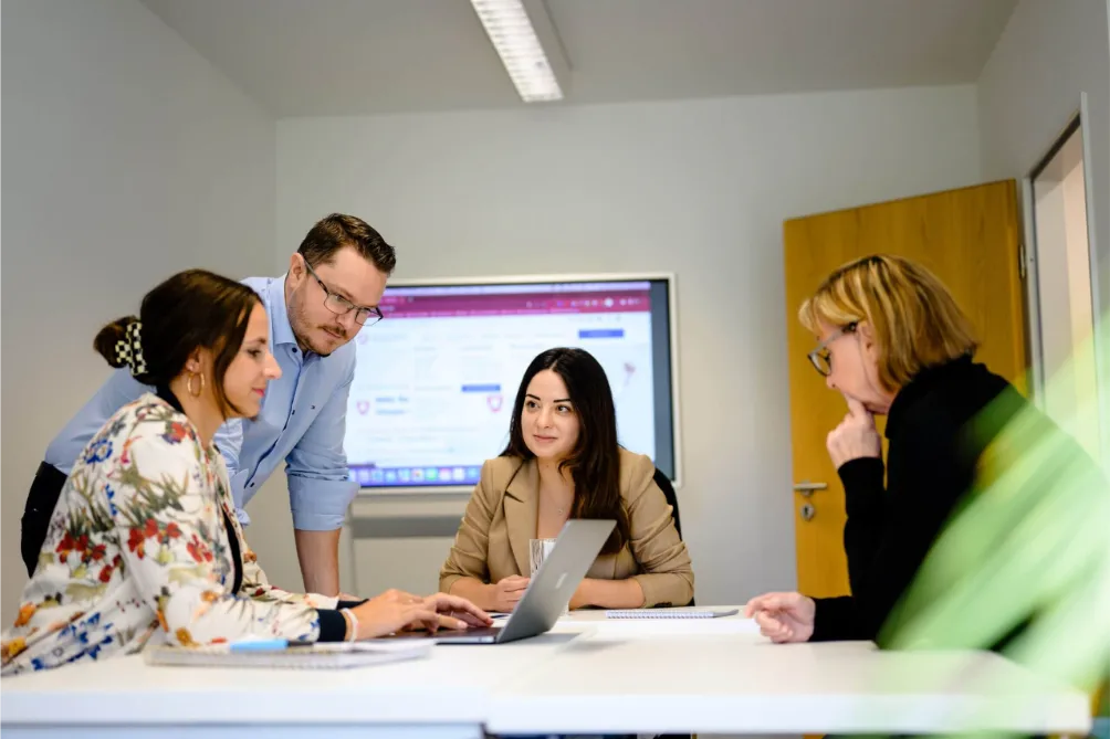 Vier Personen arbeiten im Büro gemeinsam an einem Laptop und entwickeln ein SEO-Konzept, während eine Website im Hintergrund angezeigt wird.