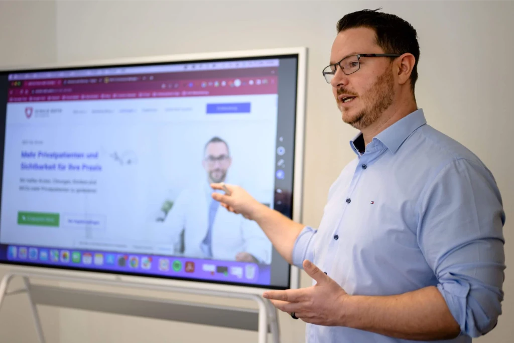 Mann mit Brille und blauem Hemd zeigt auf Bildschirm mit SEO-für-Ärzte-Website; moderner Raum, Arztfoto sichtbar.