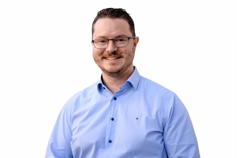 Mann mit braunen Haaren und Brille, im blauen Hemd, lächelt vor grauem Hintergrund – bereit für SEO-Thema.
