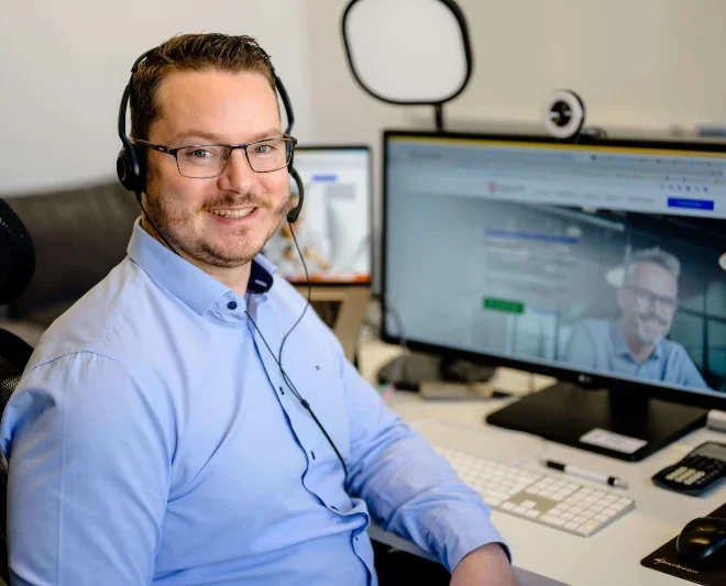 Mann mit Brille und Headset lächelt am Schreibtisch im modernen Büro einer SEO-Agentur Köln vor Computer.