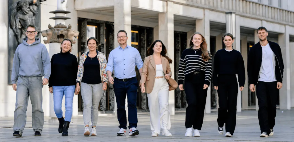 Acht unterschiedliche Menschen gehen gemeinsam im Freien spazieren, lächelnd und in legerer und professioneller Kleidung, was auf ein freundliches Team oder eine freundliche Gruppe schließen lässt.