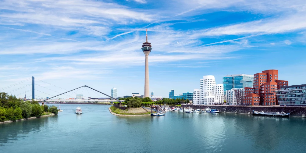 Düsseldorfer Stadtbild mit Rhein, Rheinturm, moderner Brücke und zeitgenössischen Gebäuden unter einem blauen Himmel mit dünnen Wolken.