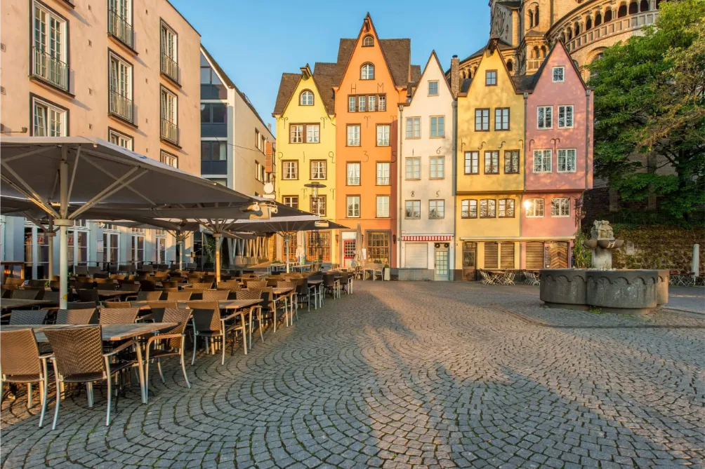 Kopfsteinpflasterplatz in Hürth mit leeren Cafétischen, farbenfrohen historischen Gebäuden und einem Steinbrunnen bei Sonnenaufgang.