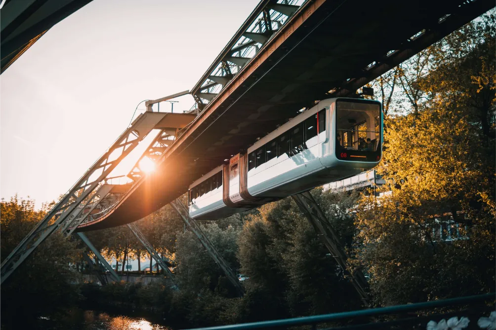 Eine Einschienenbahn gleitet bei Sonnenuntergang über einen Fluss in Wuppertal, Sonnenlicht erhellt die Szene.