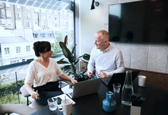 Zwei Personen unterhalten sich an einem Tisch mit Laptops im Büro einer modernen SEO-Agentur in Köln. Durch die Fenster ist die Stadtlandschaft zu sehen.