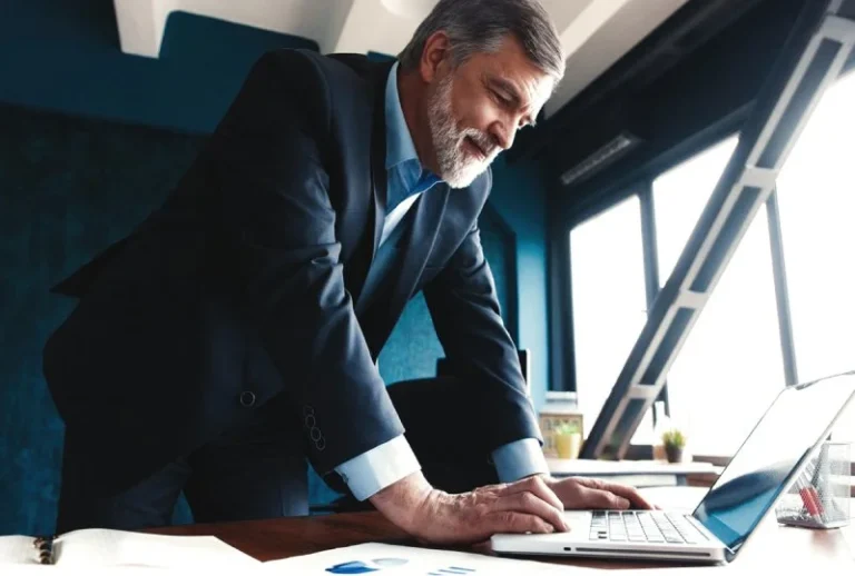 Ein älterer Mann im Anzug lächelt, während er an einem Laptop arbeitet und in einem modernen Büro eine Website für einen Steuerberater gestaltet.