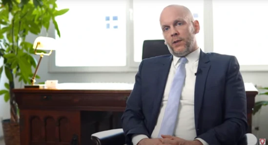 Mann im Anzug sitzt am Schreibtisch vor großen Fenstern und Pflanzen; professionelle Büroumgebung; neutraler Gesichtsausdruck.